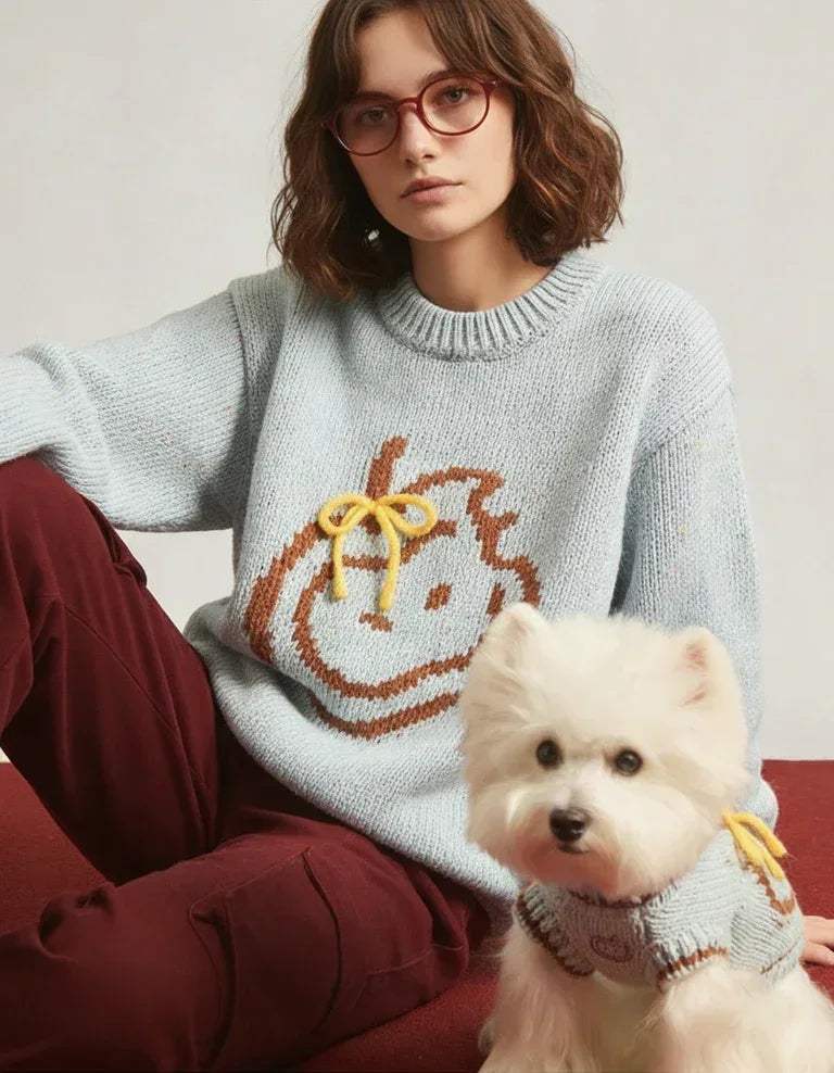 Person wearing a sweater, sitting next to a small white dog in a matching sweater.