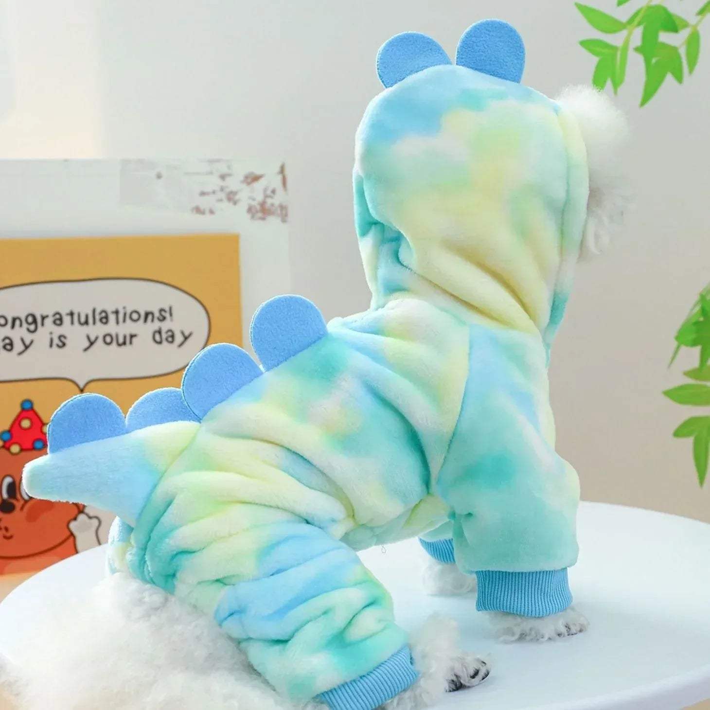 Small dog wearing a blue and green tie-dye onesie with hood on a light background.