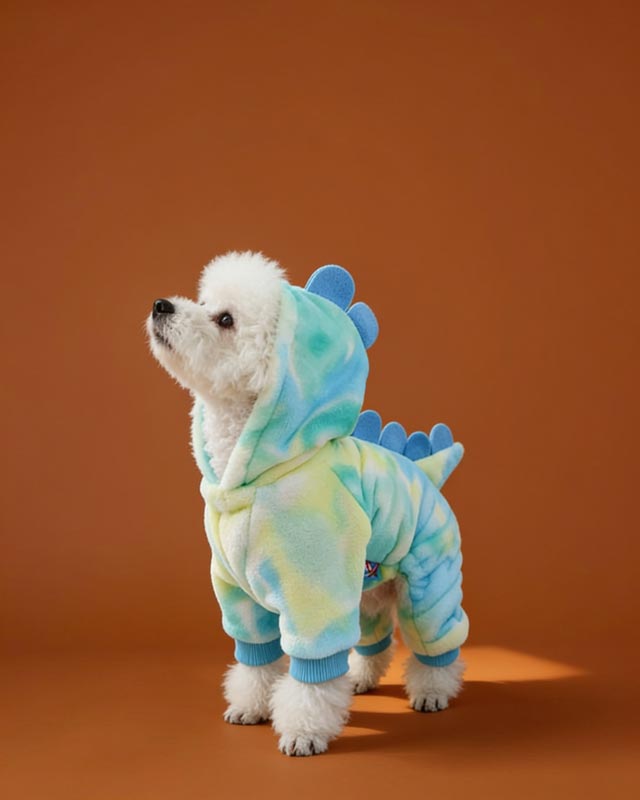 Small dog wearing a blue and green tie-dye onesie with hood on a light background.