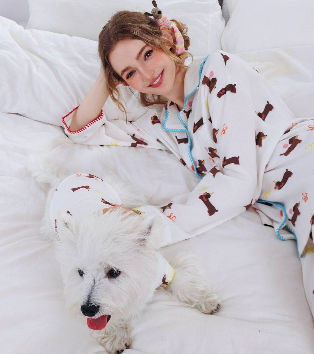 Woman in pajama set with dog pattern, lying on a bed with a small white dog.