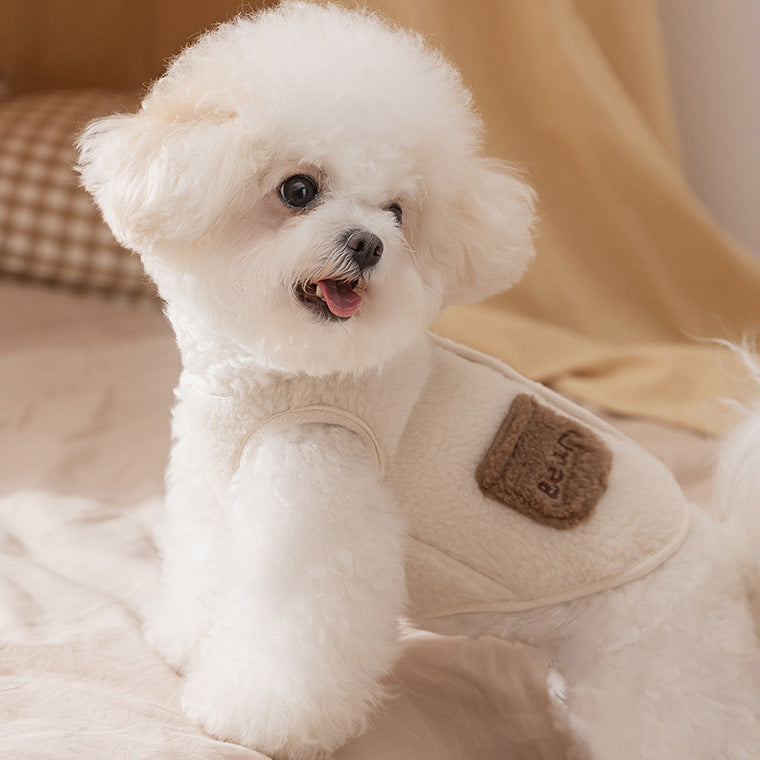Small white dog wearing a beige jacket with a brown patch on a soft surface