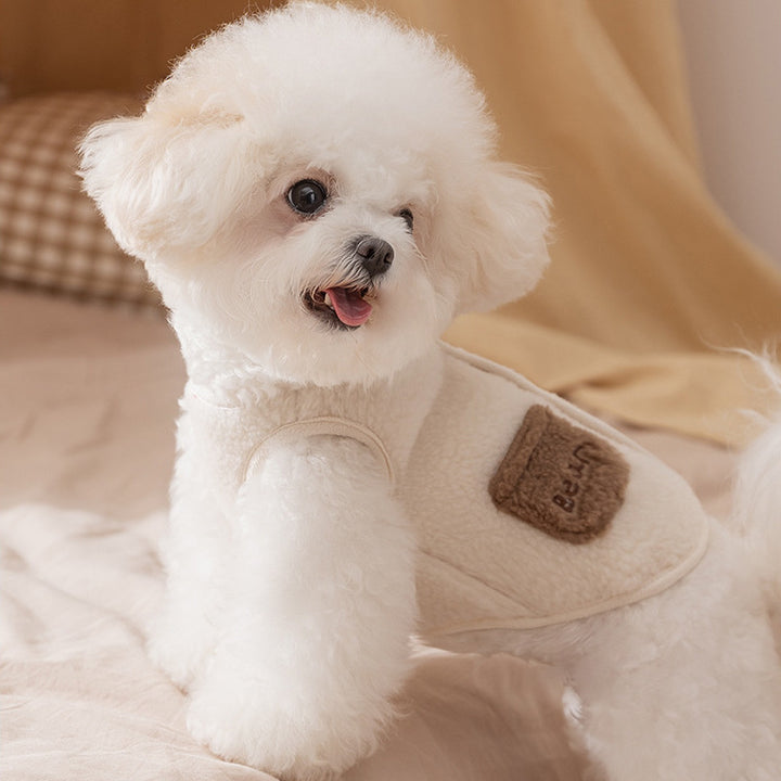 Small white dog wearing a beige jacket with a brown patch on a soft surface