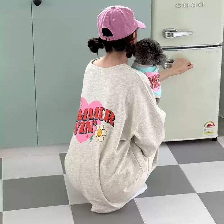 Person wearing a 'Summer Lovin' shirt and pink cap with a small dog in a carrier, standing in a kitchen.